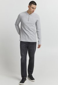 Graues Langarmoberteil mit einem Viertelreißverschluss, strukturiertem Stoff und klassischem Kragen. Kombiniert mit dunkelblauen Jeans und schwarzen Sneakers.