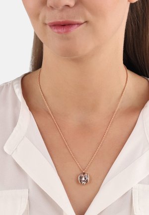 Collana in oro rosa con ciondolo ovale e pietra trasparente, incastonata in una cornice metallica, indossata sopra una blusa bianca.