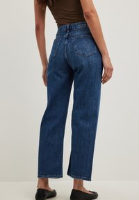 Mujer con jeans azules de pierna recta de tiro alto y zapatos planos negros, de pie con la espalda hacia la cámara contra un fondo sencillo.