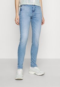 Jeans ajustados de color azul claro, hechos de denim, con una cintura de altura media y detalles de cremallera lateral, combinados con zapatos deportivos de tonos claros.
