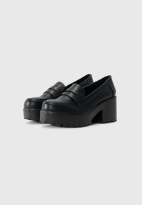 Loafers pretos em couro com plataforma, com um salto robusto e sola texturizada, apresentando um detalhe de tira costurada na parte da frente.
