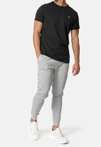 Mann trägt ein schwarzes T-Shirt mit kurzen Ärmeln, hellgraue Jogginghosen und weiße Turnschuhe. Er steht mit einer Hand in der Tasche.