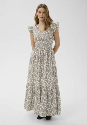 Femme aux longs cheveux portant une robe maxi blanche à fleurs avec des manches à volants et des chaussures noires, debout devant un fond uni.