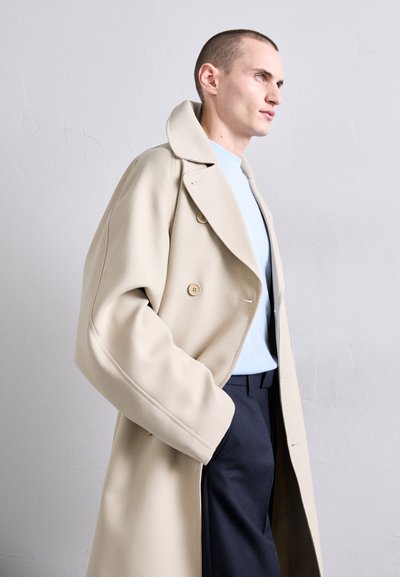 Cappotto beige doppiopetto con maniche over, ampi revers e bottoni decorativi, abbinato a un maglione azzurro chiaro e pantaloni scuri.