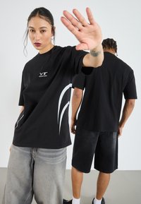 Svart t-shirt med kortaärmar som har en vit logotyp och böjda designelement. Modellen bär lösa gråa jeans, medan en andra person syns i svarta shorts.