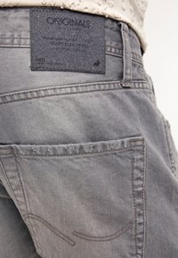 Pantalones de mezclilla gris con una etiqueta negra texturizada que dice "ORIGINALS" en la parte posterior. Presenta bolsillos cosidos y detalles sutiles.
