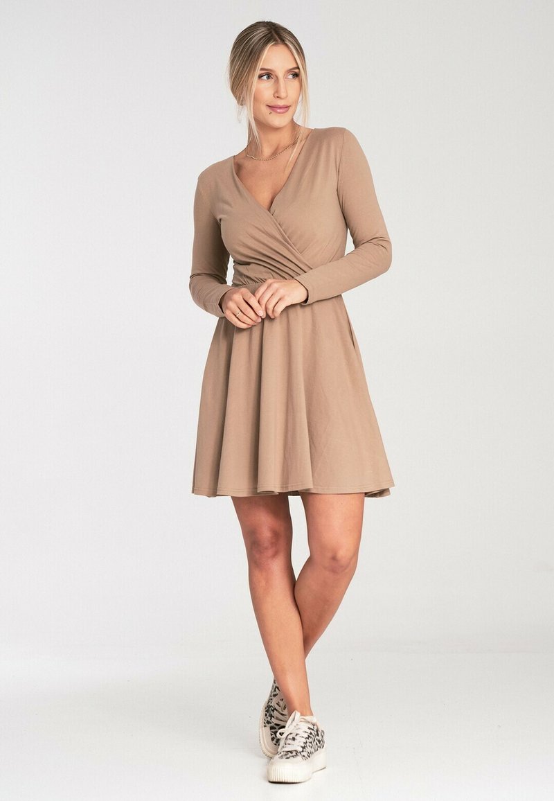 Beżowa sukienka z długim rękawem, z fasonem kopertowym i rozkloszowanym dołem. Miękki, gładki materiał. Połączona z białymi sneakersami z czarnym wzorem.