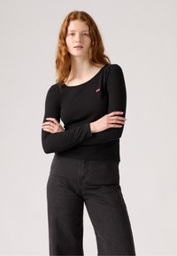 Svart langermet topp laget av mykt materiale, med rund hals og liten rød logo-lapp på brystet, kombinert med mørke jeans.