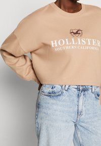 Beige zkrácená mikina s bílým nápisem "HOLLISTER SOUTHERN CALIFORNIA" a logem. Nosit s světle modrými džíny s vysokým pasem.