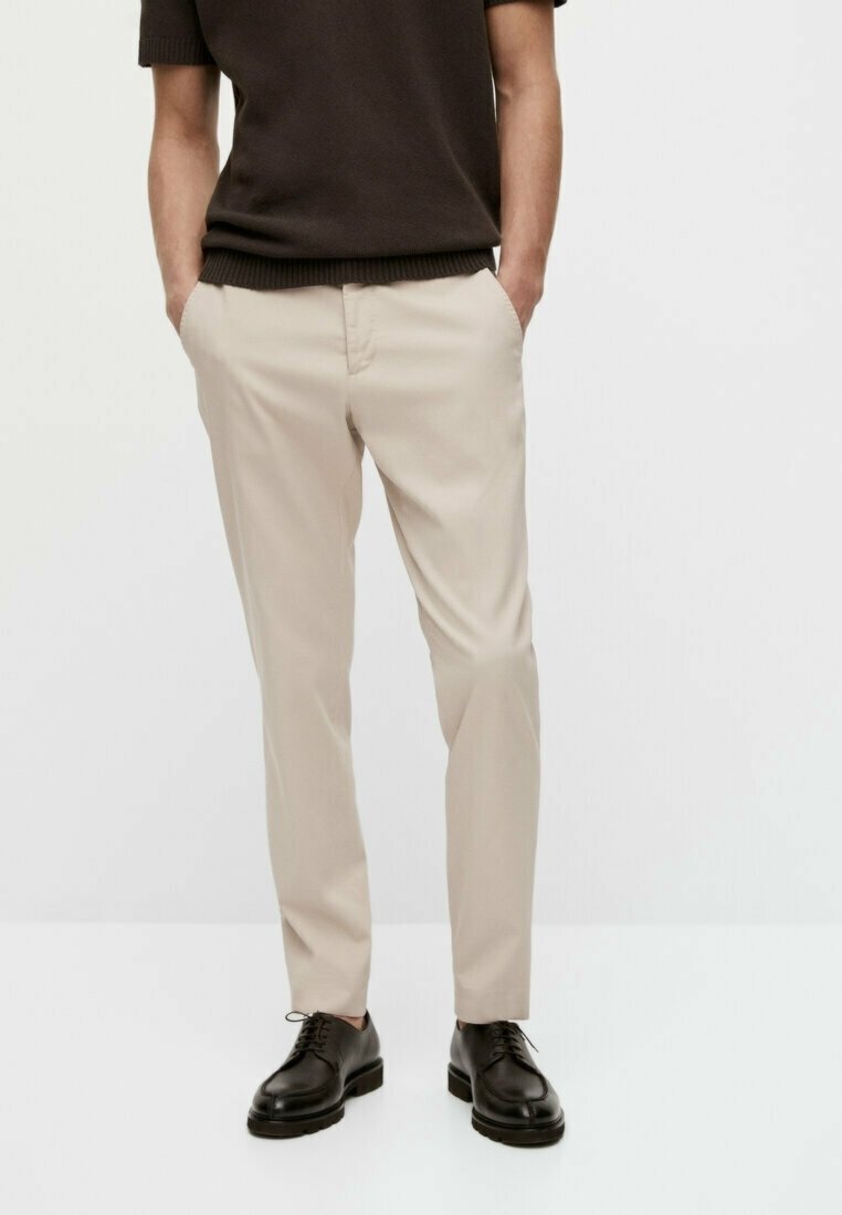 Beige chino's met een slim fit, voorzien van een platte voorkant en zijzakken, gecombineerd met zwarte nette schoenen, tegen een lichte achtergrond.