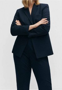 Blazer structuré bleu marine avec une fermeture à un bouton et un col cranté, associé à un pantalon fuselé assorti, tous deux en tissu lisse.