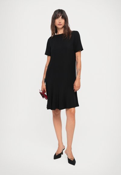 Black short-sleeve dress with a straight cut and flared hem. Worn with pointed black heels and holding a small red clutch. Smooth texture.
