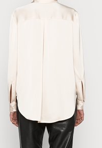 Blouse en soie crème avec des manches longues, fentes sur les côtés, ourlet arrondi et poignets boutonnés. Texture lisse et design minimaliste.