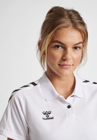 Maglietta polo bianca a maniche corte, con logo nero e strisce nere sulle spalle. Tessuto liscio con colletto classico e bottoni.