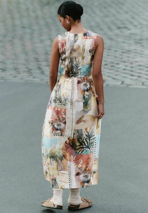 Woman with dark hair in a bun wears a sleeveless, patterned midi dress with floral and abstract designs, beige socks, and flat shoes on a paved street.