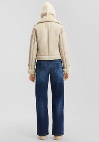 Persoon in een beige schapenvachtjack met capuchon, blauwe rechte jeans en beige fuzzy slippers, van achteren gezien tegen een effen achtergrond.