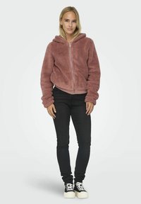 Frau, die vor einem weißen Hintergrund steht und eine staubige rosa Reißverschluss-Fleecejacke, schwarze Skinny-Jeans und schwarz-weiße Sneakers trägt.