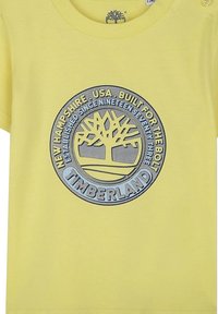 Gele katoenen t-shirt met een ronde, grijze logo en een boomontwerp, inclusief tekstelementen zoals "Timberland" en het oprichtingsjaar.
