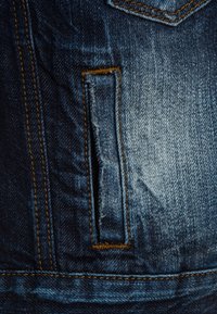 Dark blue denim fabric with a distressed pocket, featuring contrasting orange stitching. The texture is smooth with faded areas.