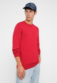 Pull en tricot rouge à manches longues et poignets côtelés, assorti à un jean en denim bleu clair et une casquette bleu foncé. Couleur unie, design simple.