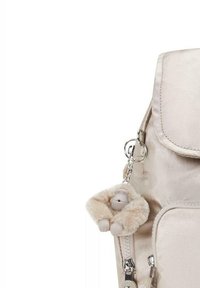 Sac à dos beige métallique avec fermeture à glissière, rabat avant et porte-clés avec un accent de singe en peluche. Texture de tissu lisse.