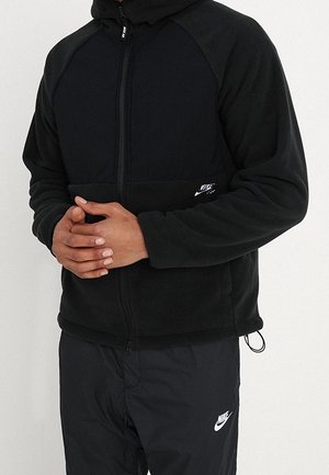 Mand iført sort Nike lynlåstrøje og sorte Nike bukser med hænderne foldet foran, stående mod en hvid baggrund.