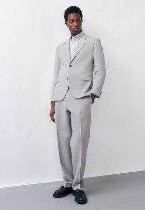 Άνδρας όρθιος, φοράει ανοιχτογκρι κοστούμι με σακάκι δύο κουμπιών και αντίστοιχο παντελόνι, λευκό πουκάμισο και μαύρα παπούτσια, πάνω σε απλό λευκό φόντο.