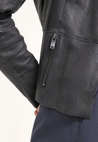 Giacca nera in finta pelle con un motivo testurizzato a pelle di serpente. Presenta una cerniera laterale e un design aderente. Finitura liscia e lucida.