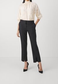 Kvinna i en krämfärgad mönstrad blus med puffärmar och svarta skräddarsydda byxor i ankellängd med framslitsar samt svarta högklackade skor.