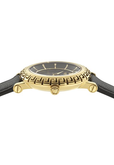 Púzdro hodiniek v zlatej farbe s lunetou s motívom gréckeho kľúča, čierny ciferník so zlatými značkami, korunka s hlavou Medúzy a čierny kožený remienok.