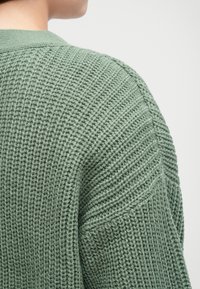 Strikken grønn cardigan viser et teksturert, ribbet design med en avslappet passform. Synlige sømdetaljer og en strukturert krage.