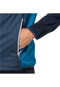 Jacka för idrott med dragkedja i marinblått och teal-färgade paneler, texturerad mörk ärm och hand i fickan, som visar på funktionella designelement.