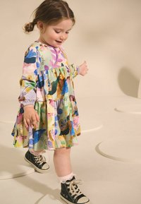 Robe florale multicolore à manches longues, avec un design en étages et des poignets élastiques. Portée avec des chaussures montantes noires et des chaussettes blanches en dentelle.