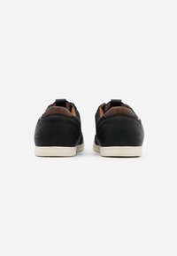 Baskets noires avec une texture en cuir lisse, accents marron contrastants et semelles en caoutchouc crème. Possède une languette pour un enfilage facile.