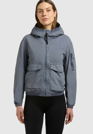 Mujer vistiendo una chaqueta gris con capucha y cremallera, con bolsillos delanteros con solapa y un bolsillo con cremallera en la manga, combinada con pantalones negros.