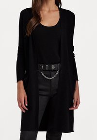 Svart cardigan med öppen front, långärmad, ribbad struktur och två framfickor, kombinerad med ett svart linne och jeans. Detalj med silverkedjebälte.