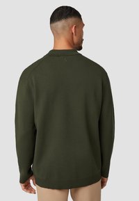 Olijfgroene gebreide trui met lange mouwen, geribbelde manchetten en zoom. Bevat een subtiele logo nabij de kraag. Zachte textuur, relaxte pasvorm.