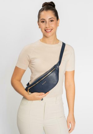 Bolso cruzado de cuero azul marino con una superficie texturizada, forma curva, cremallera dorada y correa ajustable, sostenido contra un fondo claro.