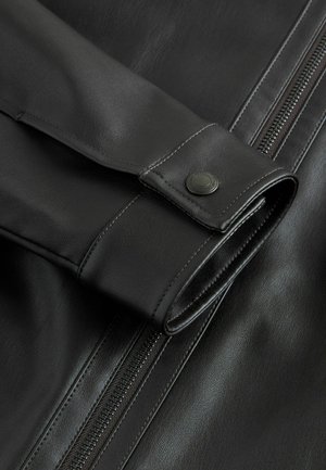Nahaufnahme einer schwarzen Lederjacke mit glatter Textur, einem Knopfakzent am Ärmel und einem sichtbaren Reißverschluss an der Seite.