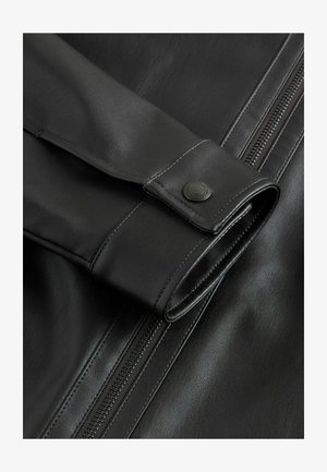 Primer plano de una chaqueta de cuero negra que presenta una textura suave, un acento de botón en la manga y una cremallera visible a lo largo del costado.