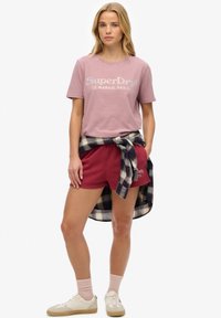 Lyserød bomulds t-shirt med "SuperDry" logo, bordeaux shorts og enternet skjorte bundet om taljen. Modellen har lyserøde sokker og beige sneakers på.