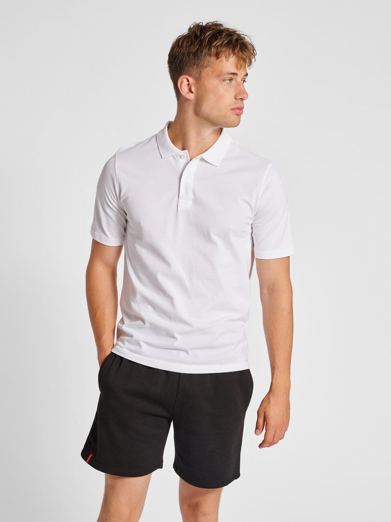 Junger Mann, der ein weißes kurzärmeliges Poloshirt und schwarze Shorts trägt, steht mit den Händen in den Taschen und schaut nach rechts.