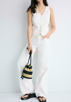 Donna che indossa un gilet bianco senza maniche con bottoni e pantaloni a gamba larga, che tiene una borsa a coulisse a righe nere e beige.