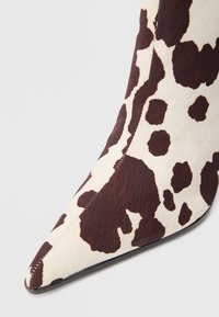 Botine cu vârf ascuțit, confecționate din piele de bovină, cu un model lucios de imprimeu de vacă în nuanțe de maro și crem. Textură netedă și design elegant.