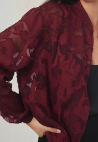 Cardigan transparent bordeaux avec broderie florale, motifs texturés et coupe décontractée. Tissu doux avec une apparence légère.