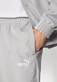 Grå sportsjakke med en glat tekstur, der har elastiske manchetter og et hvidt Puma-logo på venstre bryst. Hånden er i en lomme.