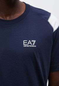 Άτομο με σκουρόχρωμο δέρμα που φοράει μπλε ναυτικό t-shirt Emporio Armani EA7 με λευκή ρίγα στον ώμο, κοντινό πλάνο στο στήθος και τον λαιμό.