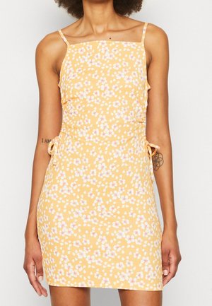 Mujer con vestido corto sin mangas amarillo con estampado floral blanco y rosa y tirantes finos.