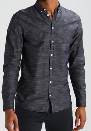 Mand iført en tætsiddende, mørkegrå skjorte med subtile vandrette striber, kombineret med sorte bukser, stående med arme afslappet ned langs siden.