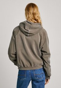 Sudadera con capucha en un verde olivo suave, con bordes acanalados, textura blanda y corte relajado. Combinada con jeans de mezclilla azul.